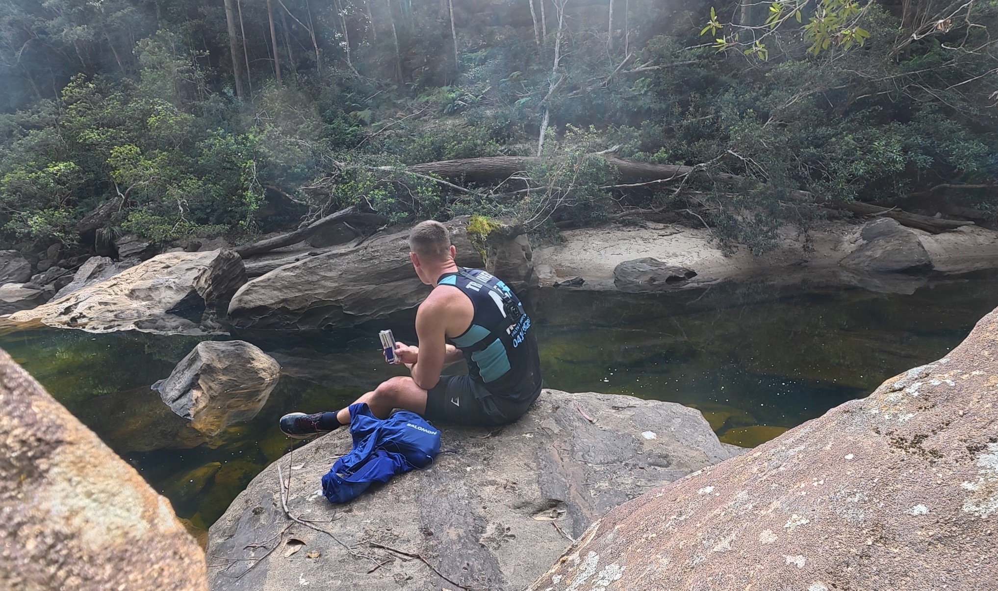 Nathan sitting on a boulder beside a pool on the Grose River, checking his phone