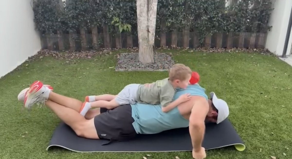 Berdo doing push ups on a mat in the backyard with his young son lying on his back